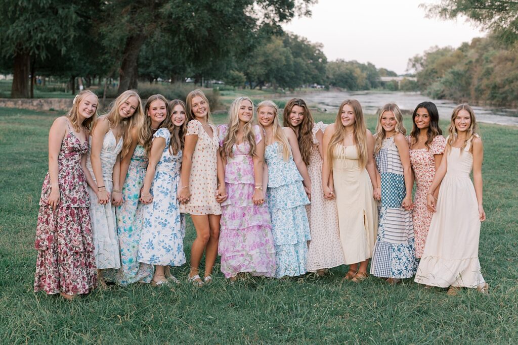 Group of high school senior girls rep team posed casually holding hands at park in floral dresses.
