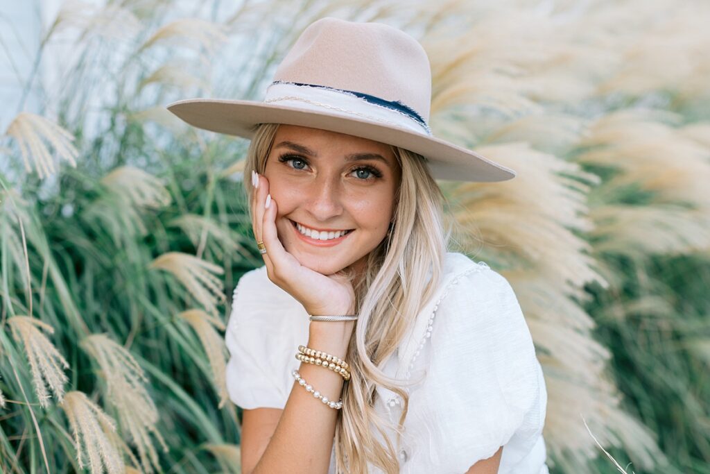 Close up senior portrait of girl with relaxed, natural smile wearing a white dress and cute hat in a field