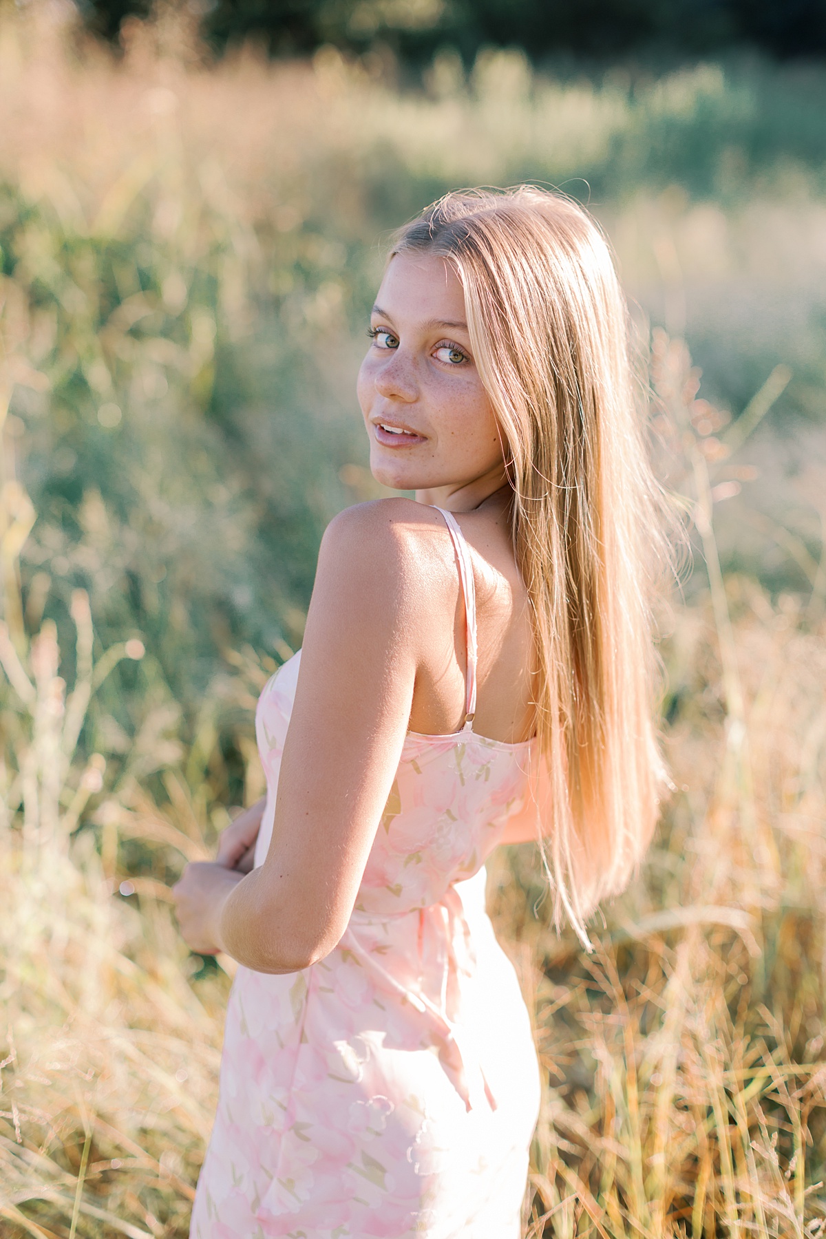 Golden hour Malibu senior photo of girl looking over shoulder in a pink dress in a sunlit field.
