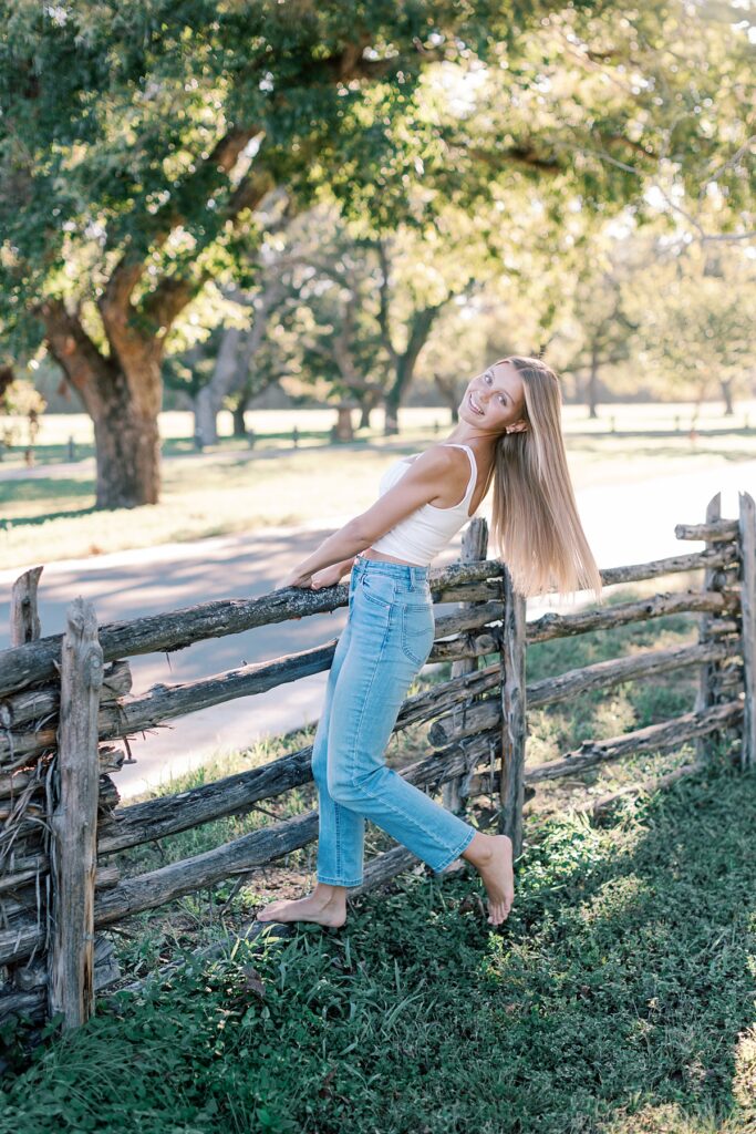 Southern California senior picture idea with girl barefoot smiling and leaning back on wooden fence in a sunlit field.
