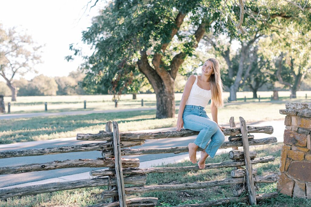 High school senior girl sitting barefoot on a wooden fence in a grassy Malibu field, captured during golden hour.