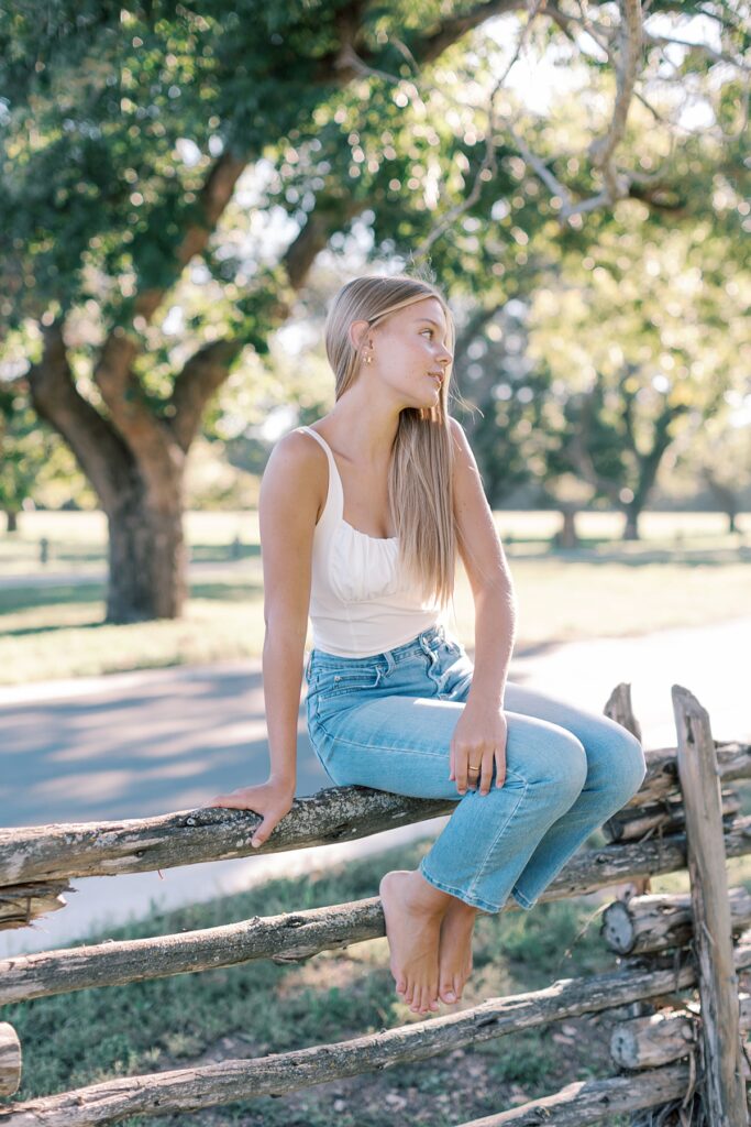 Southern California outdoor senior portrait idea of a girl in jeans sitting on rustic fence under tree.
