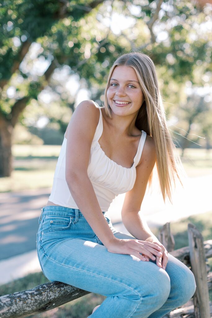 Smiling senior girl captured at golden hour in a Malibu field, sitting on a rustic fence for outdoor senior photo inspiration.