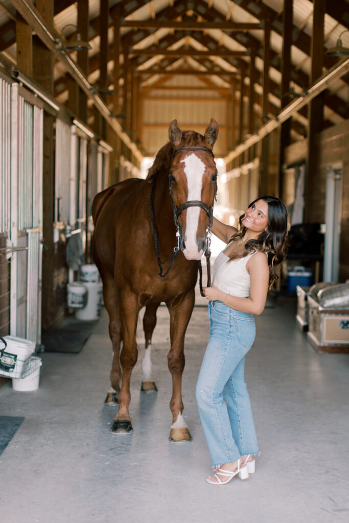 Senior girl smiling next to her horse inside barn aisle – stylish senior portraits with equestrian theme.