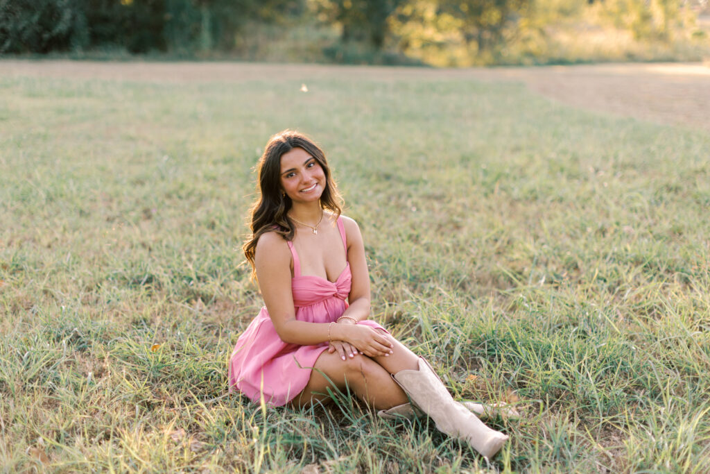 Senior girl poses sitting in soft sunset light with relaxed posture and boots.