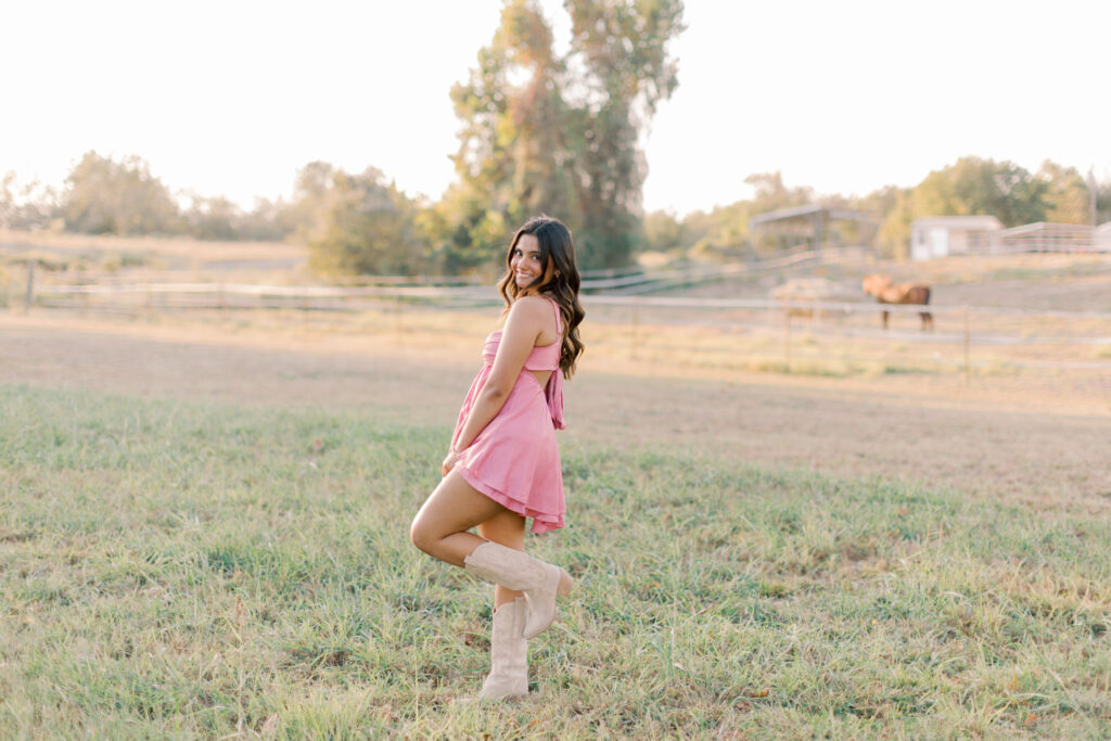 Southern California senior portrait girl smiles with her leg popped, golden grass and horse stables in background