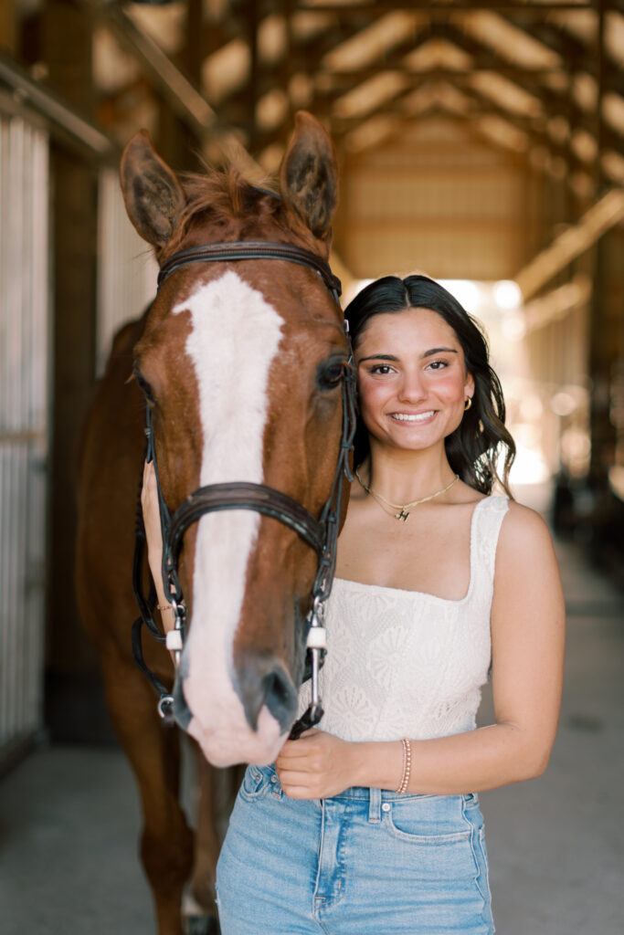 Senior girl smiling next to her horse inside barn aisle – stylish senior portraits with equestrian theme.