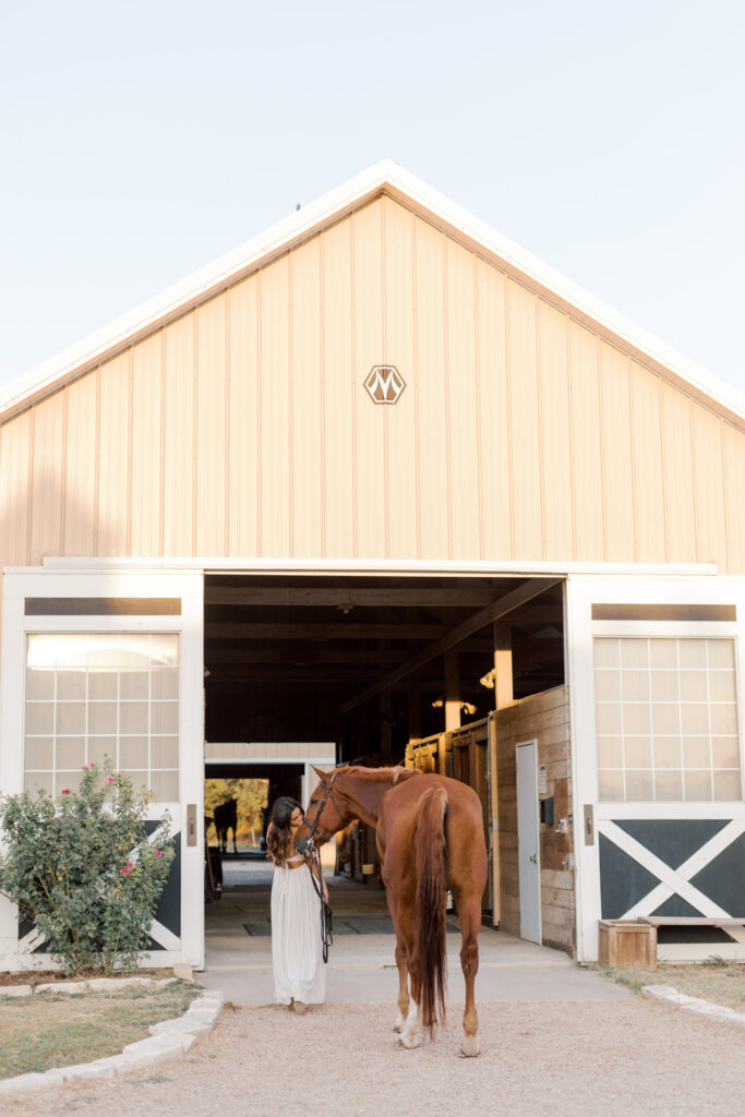 Natural light photo of a senior with her horse by a white barn – simple and authentic Southern California portrait.