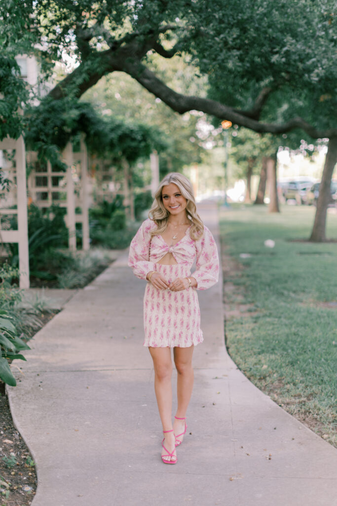 Senior girl smiling in pink floral dress walking on a tree-lined sidewalk during golden hour