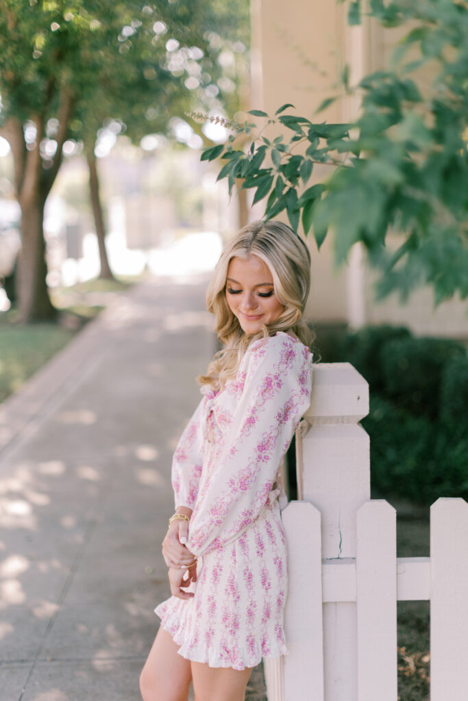 High school senior softly smiling and leaning against a white picket fence in sunlight