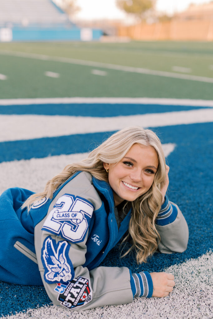 Blonde girl in soccer uniform laying on center field logo with ball, senior photos Southern California