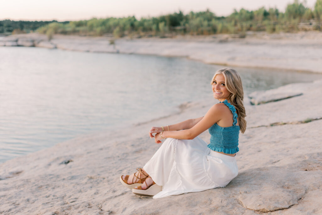 Blonde senior smiles over her shoulder near the water in a blue ruffle top. Southern California senior portraits with soft, natural light.