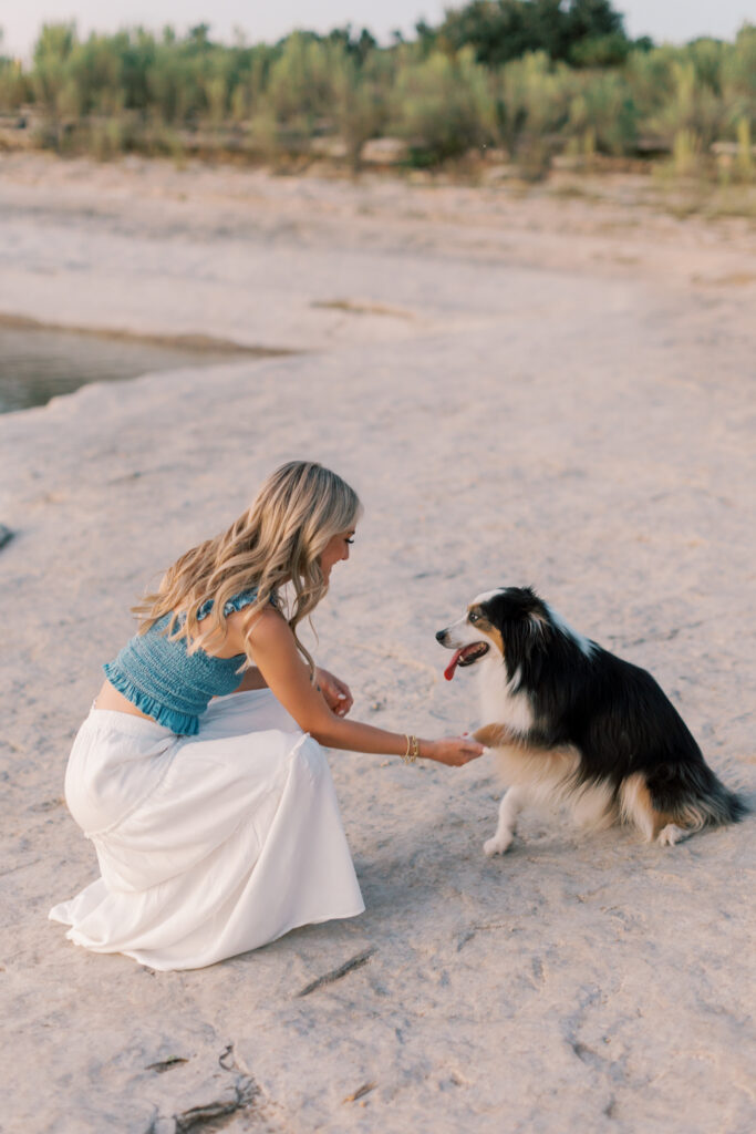 Senior girl crouches to shake hands with her dog on a rocky lakeside trail. Southern California senior photographer for natural, candid portraits.