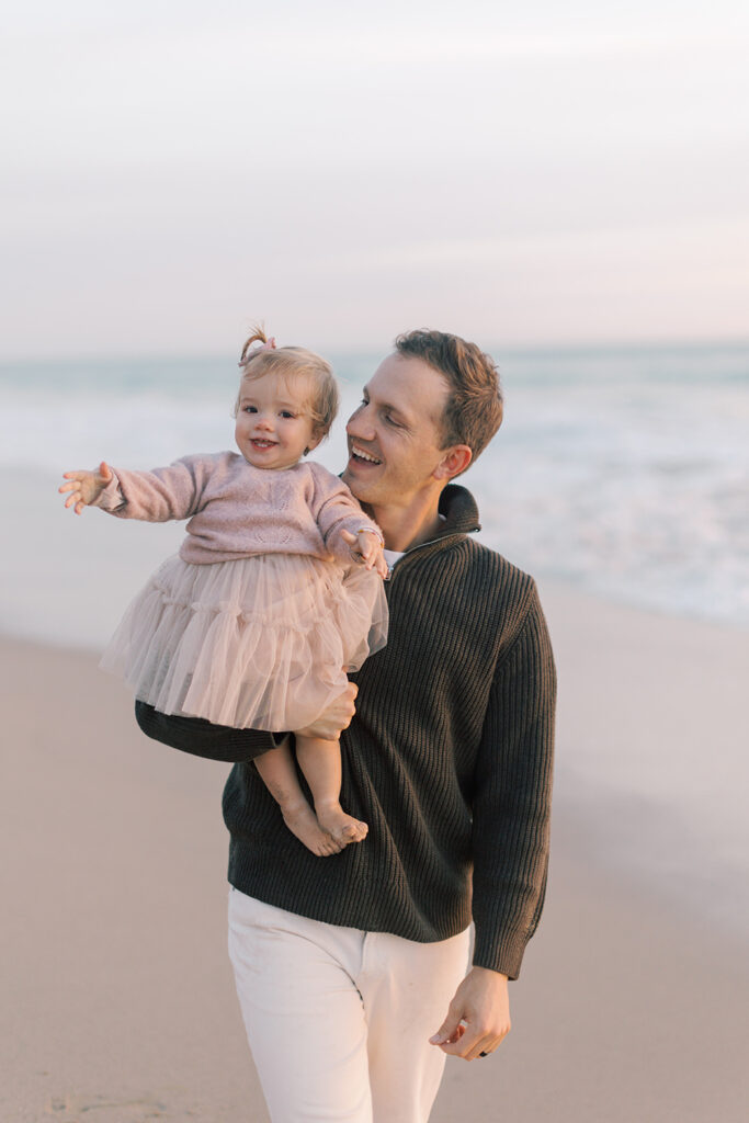 Dad holding baby girl smiling for family portrait at Westward beach in Malibu.