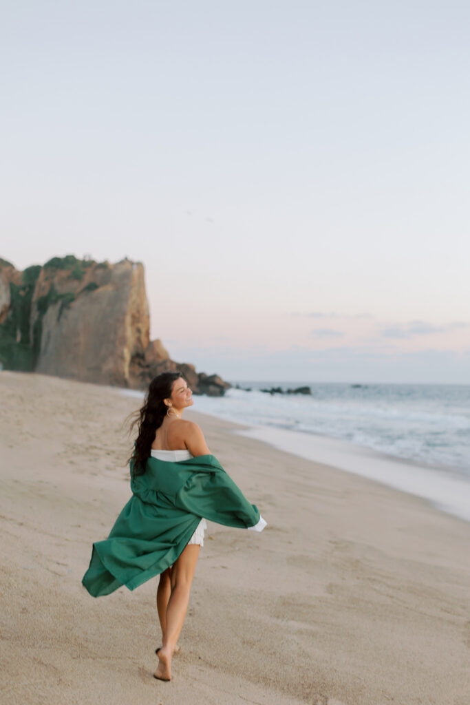 high school senior walking along beach at Point Dume Malibu in cap and gown with ocean beside her, candid graduation photos Malibu senior photographer