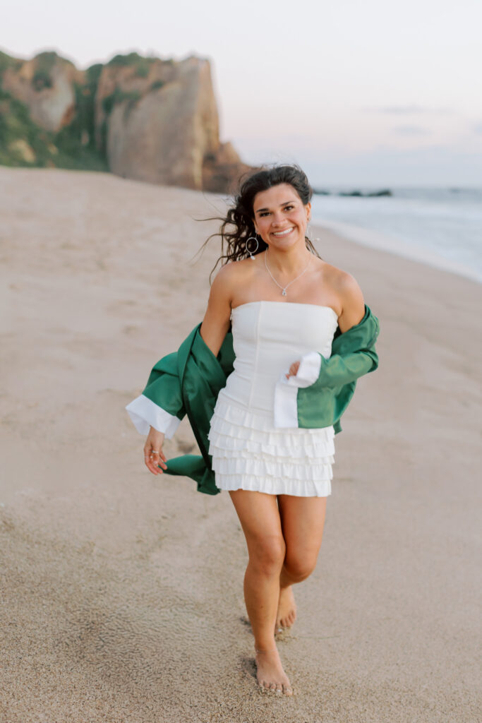 full body photo of Thousand Oaks High School senior walking on beach at Point Dume Malibu wearing cap and gown with ocean waves, graduation session Malibu senior photographer