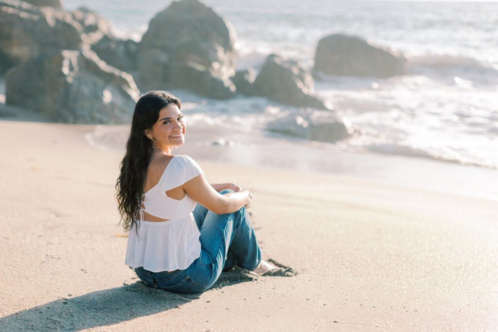 senior girl sitting in the sand on Malibu beach with legs turned to the side, looking back over her shoulder with ocean waves behind her