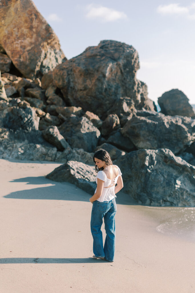 high school senior girl standing barefoot on Malibu beach near large rocks at Point Dume, looking down with relaxed natural pose and ocean behind her