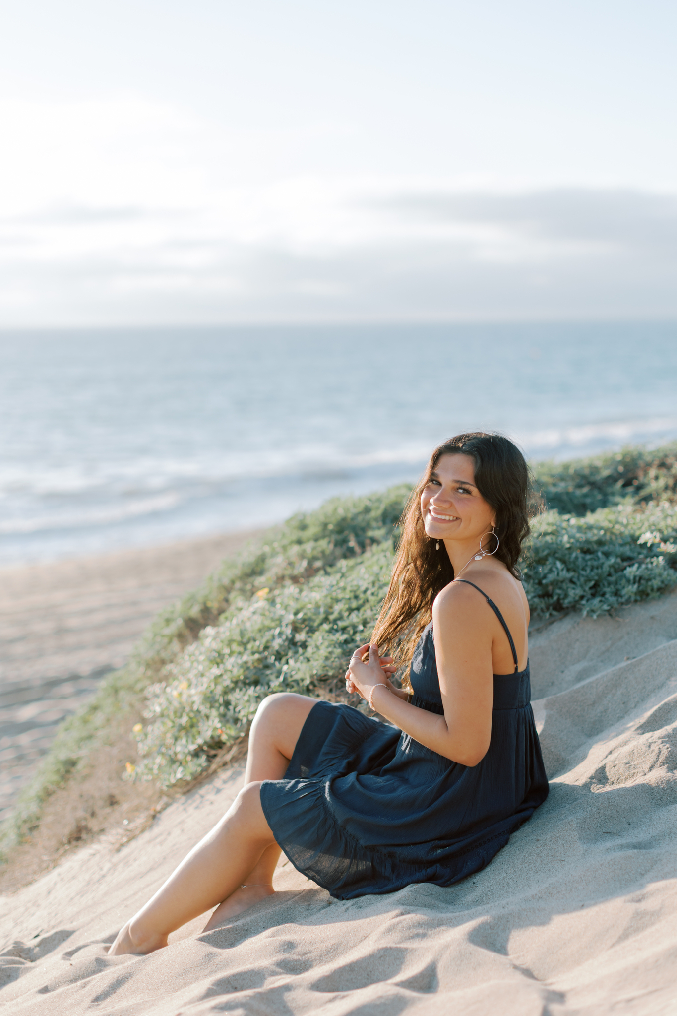 Thousand Oaks High School senior sitting on sand at Point Dume Malibu wearing a navy dress, smiling toward the camera during golden hour beach senior photos