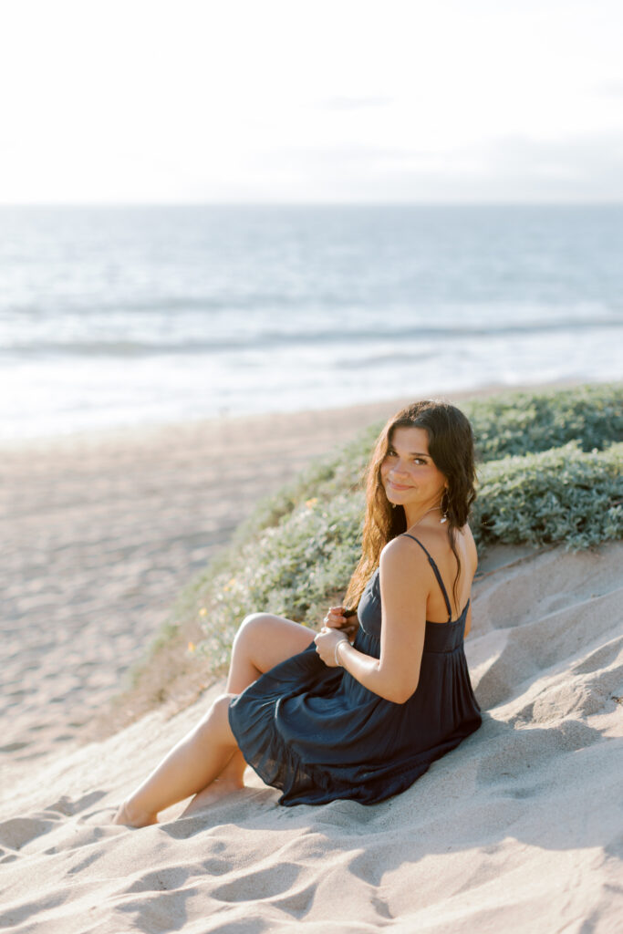high school senior sitting on sand dune at Westward Beach Malibu smiling toward camera, relaxed posing Southern California senior photographer