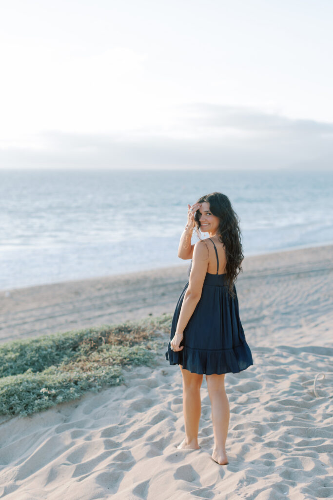 high school senior standing in sand at Westward Beach Malibu looking back over shoulder in navy dress, natural light senior photos Thousand Oaks photographer