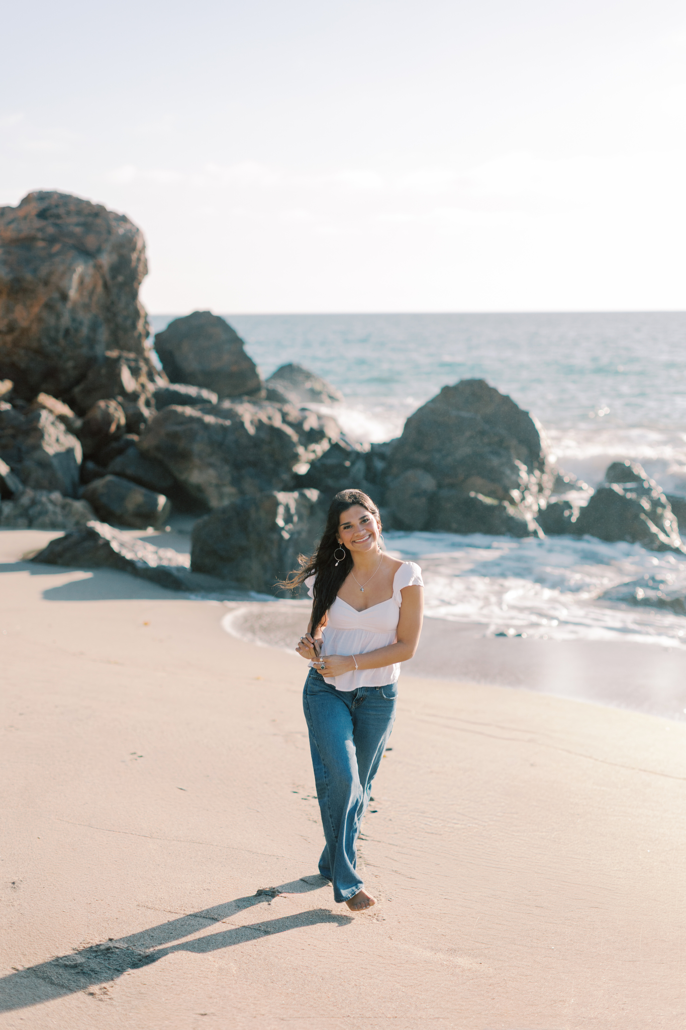 high school senior girl walking barefoot along Point Dume beach in Malibu near coastal rocks, smiling in natural golden hour light