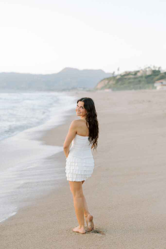 high school senior walking along shoreline at Westward Beach Malibu looking back smiling, barefoot beach portrait in white dress Southern California senior photographer