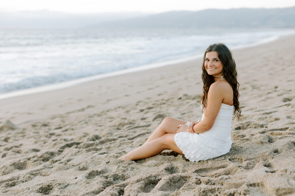 high school senior sitting in sand at Westward Beach Malibu in white dress facing camera, relaxed natural posing beach senior photos Southern California