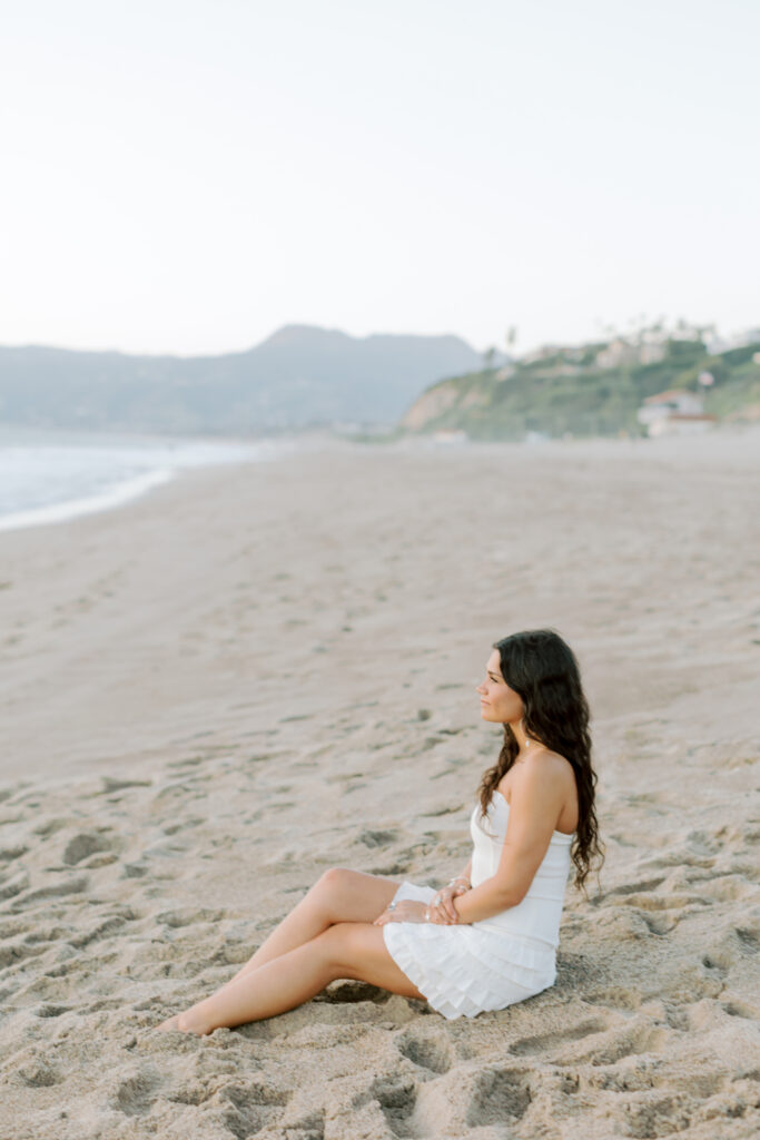 high school senior sitting on sand at Westward Beach Malibu with ocean and coastline in background, wide beach portrait Thousand Oaks senior photographer