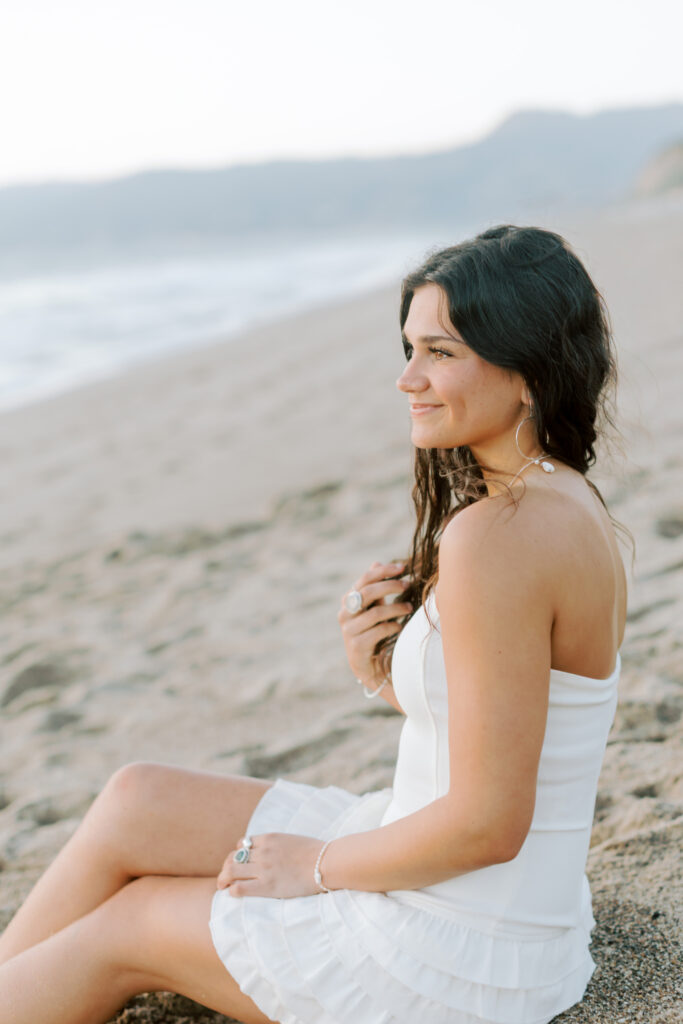 high school senior sitting on sandy beach at Point Dume Malibu looking toward ocean, side profile portrait in white dress Malibu senior photographer