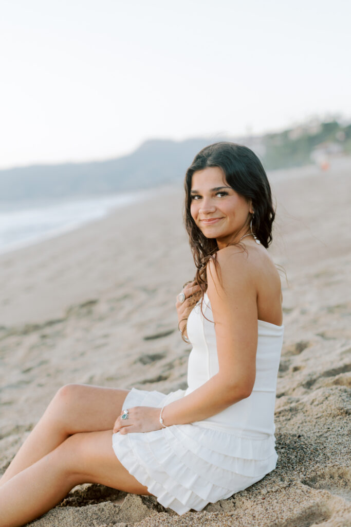 Thousand Oaks high school senior sitting in sand at Westward Beach Malibu wearing white dress, soft natural light beach senior portrait