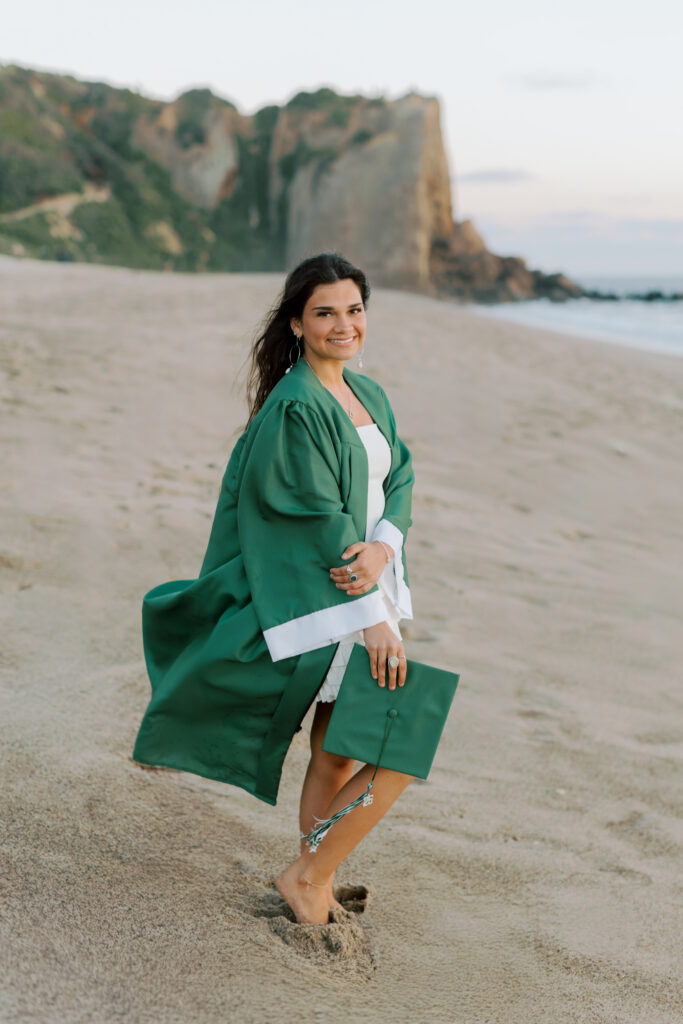 Thousand Oaks High School senior posing barefoot on beach at Point Dume Malibu wearing green cap and gown, looking back and smiling during graduation photos