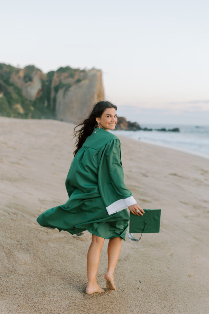 Thousand Oaks High School senior smiling in cap and gown at Point Dume Malibu beach with cliffs in background, natural light senior portraits