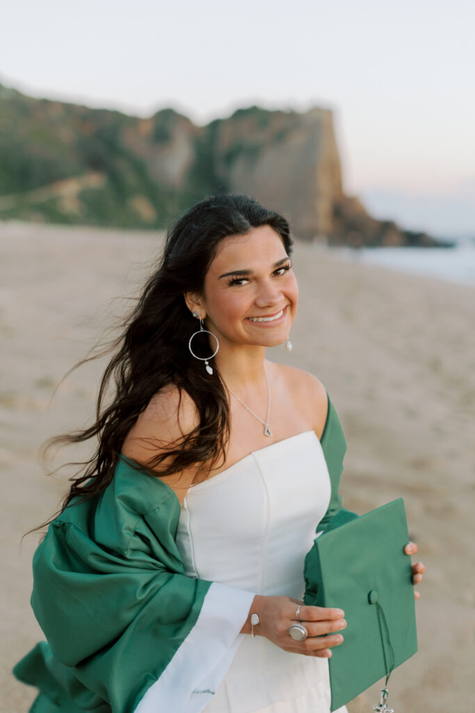 Thousand Oaks High School senior smiling in cap and gown at Point Dume Malibu beach with cliffs in background, natural light senior portraits