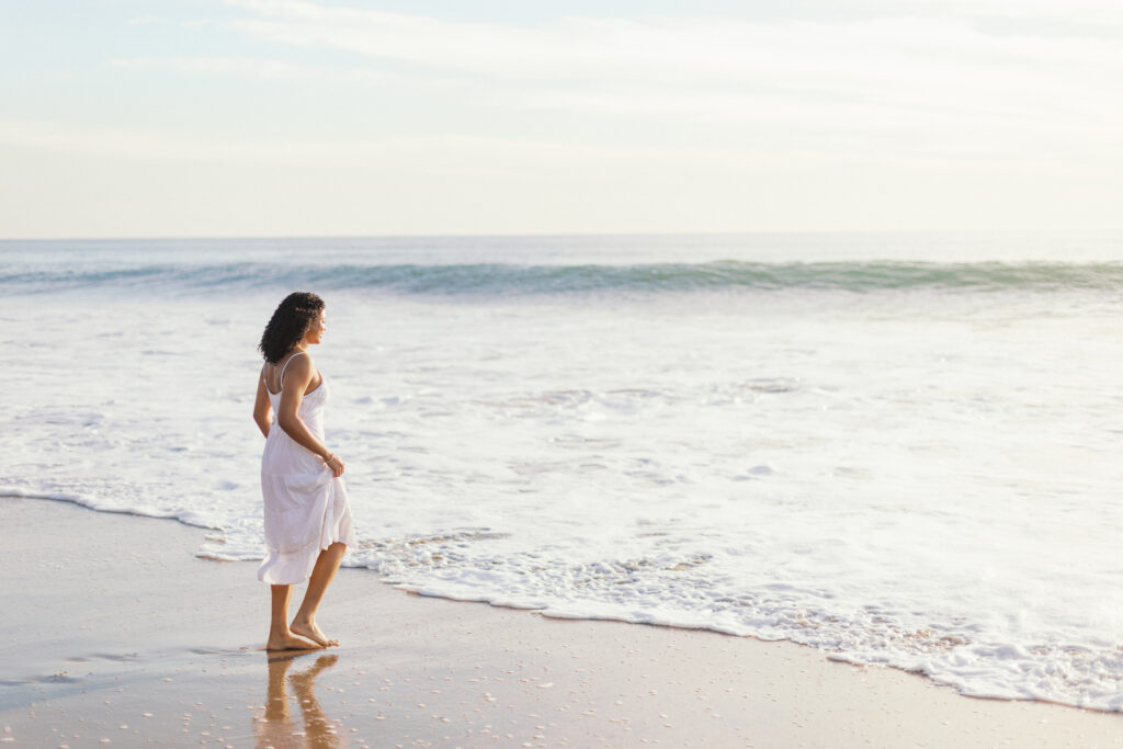 Oaks Christian senior standing at the shoreline at Westward Beach Malibu during golden hour in a white dress